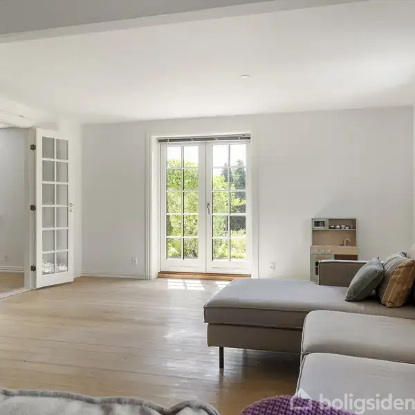 Stue med en stor, lys sofa centreret mod store glasdøre, der fører ud til en grøn have. Rummet har lyse trægulve og hvide vægge.