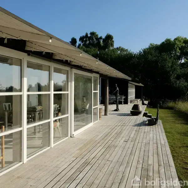 En træterrasse strækker sig foran et hus med store vinduespartier. Solen skinner, omgivet af græs og træer. Der står møbler på terrassen under en blå himmel.