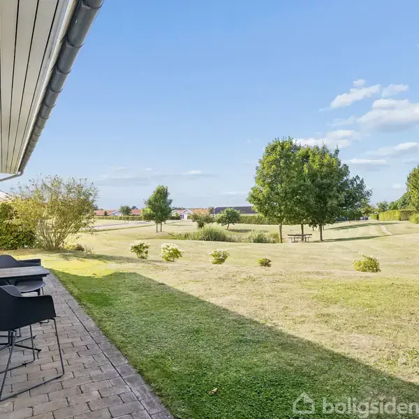 En hvid bygning med to sorte stole står stille langs en flisebelagt terrasse. I baggrunden er en velplejet græsplæne med træer og buske under en klar blå himmel.