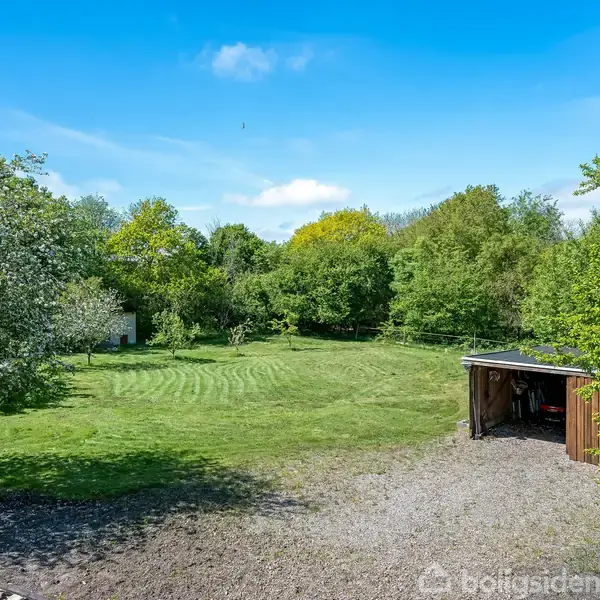 En velplejet græsplæne strækker sig gennem en stor, frodig have med tæt omgivende træer. Til højre står et lille skur ved en grusbelagt sti under en klar blå himmel.