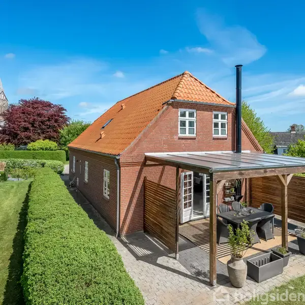 Rødt murstenshus med rødt tegltag står i en grøn have med velplejet plæne og hække. En overbygget terrasse med bord og stole ses i forgrunden, kirketårn i baggrunden.