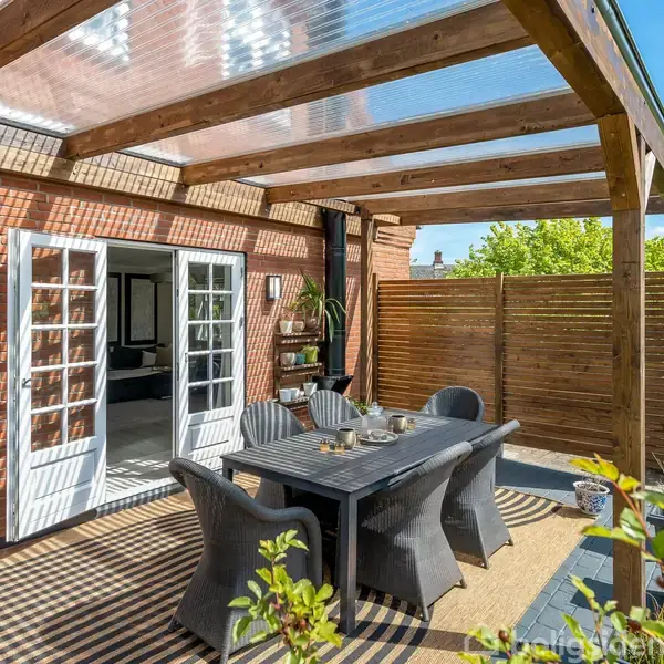 Overdækket terrasse med spisebord og stole under en trækonstruktion. Sollys filtrerer gennem taget. Omgivet af en murstensbygning, træhegn og grønne planter. Franske døre åbner til huset.