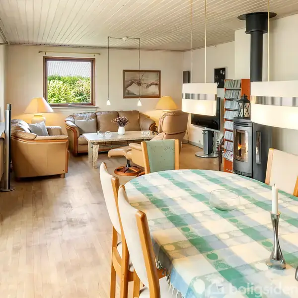 Spisebord med dug og stole står i en stue med sofaarrangement, brændeovn og lamper. Rummet har trægulve og store vinduer med udsigt til en have.