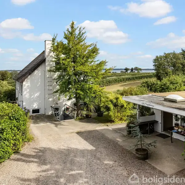 Hus med hvid facade står omgivet af frodig grønt, mens en grusvej fører forbi. Træer og buske udgør den landlige omgivelser, og himlen er klar med spredte, hvide skyer.