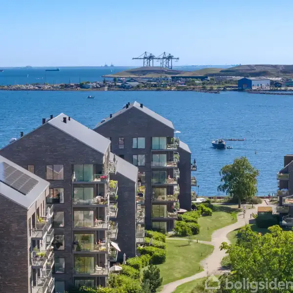 Lejlighedskomplekser med solbeskinnede altaner står tæt ved en blå havn. I baggrunden ses kraner og både mod en klar himmel. Grønne områder omkranser bygningerne.