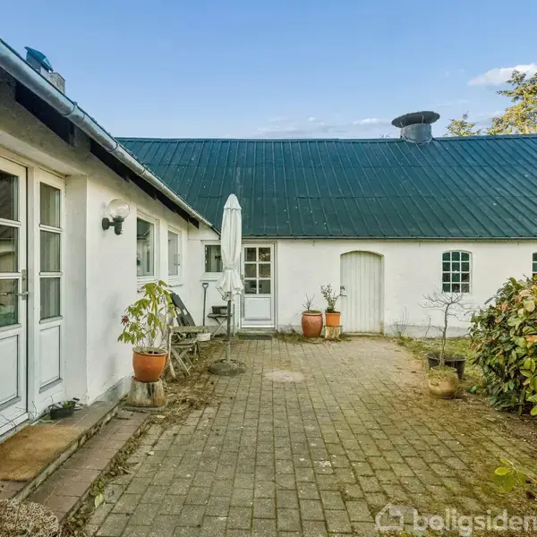 Hvidmalet hus omgivet af en brostensbelagt gårdsplads med potteplanter og havemøbler. Grønne planter og blå himmel i baggrunden.
