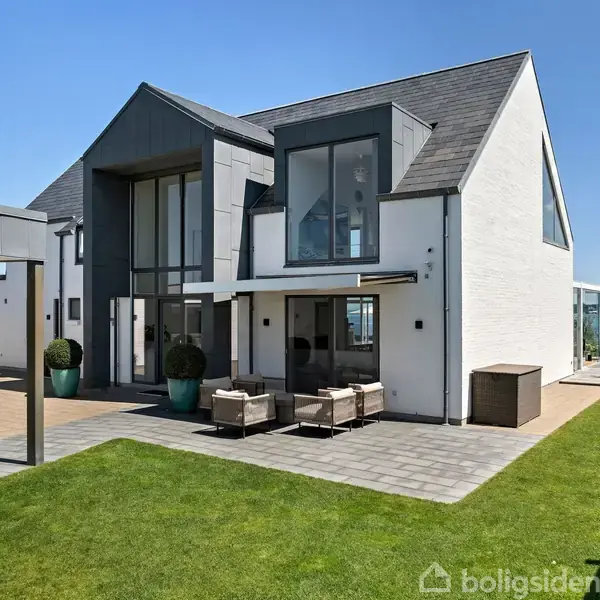 Moderne to-etagers hus står i en velplejet have ved vandet. Huset har store vinduer og en terrasse med havemøbler under en blå himmel.