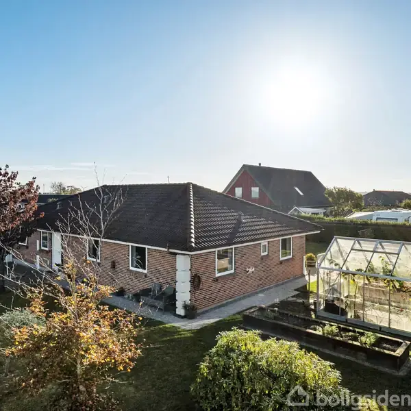 En murstenshus med sort tag står i en velplejet have. Ved siden af er et drivhus. Et dansk flag vajer på en flagstang. Solen skinner på stedet.
