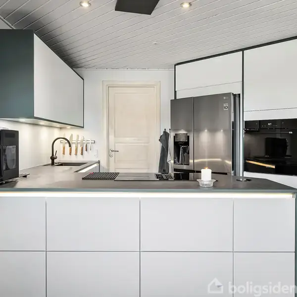 Et moderne køkken med hvide skabe og skuffer omgiver et køleskab, kaffemaskine og ovn. En dør er placeret i baggrunden, og en enkelt lysestage står på bordet.