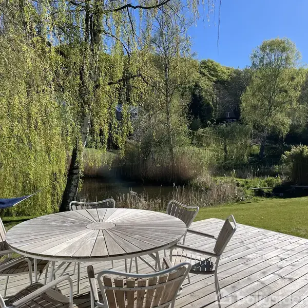Rundt havebord med stole står på en træterrasse ved en grøn have. Hængekøje hænger ved træer foran en dam med grønt område i baggrunden.
