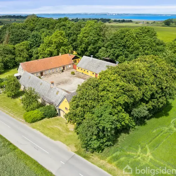 En gård med røde og grå tage omgivet af tæt bevoksede træer. Gården ligger ved en vej, og i baggrunden ses marker og en blå sø eller fjord.
