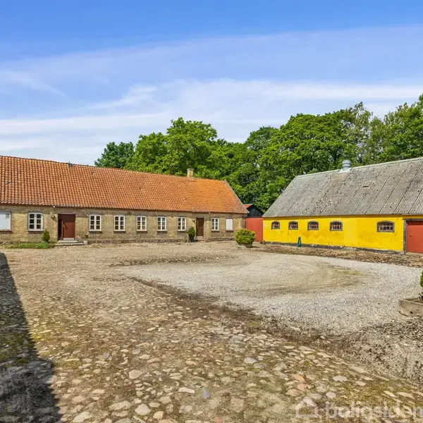 Gårdsplads omgivet af gulmalede bygninger med røde døre og klassiske tegltage. Stenbelagt gård med grusområde i midten, omkranset af grønne træer under en klar blå himmel.