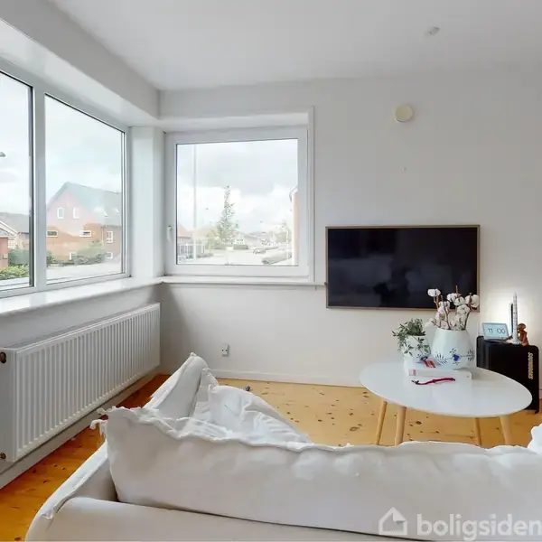 Sofa i hvid stue, står foran stort vindue med udsigt til huse. Et rundt bord og en vægmonteret fladskærm er tilstede, omgivet af enkel moderne indretning.