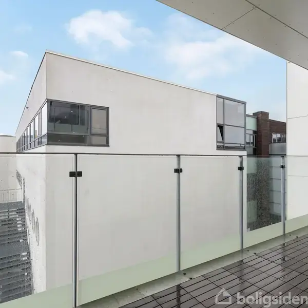 En balkon med glasrækværk står stille i en moderne bygningskompleks. Bygningens hvide facade med store vinduer ses i baggrunden under en blå himmel med få skyer.