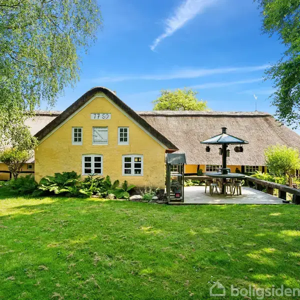 Et gult bindingsværkshus med stråtag står omgivet af en grøn græsplæne og træer. Foran huset er en terrasse med et spisebord under en parasol. Huset har teksten "1901" på gavlen.
