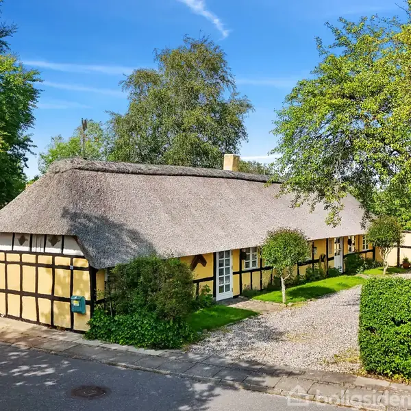 Bindingsværkshus med stråtag står stille blandt frodige grønne træer og buske i solskinsvejr. Grusvej leder op til hoveddøren.