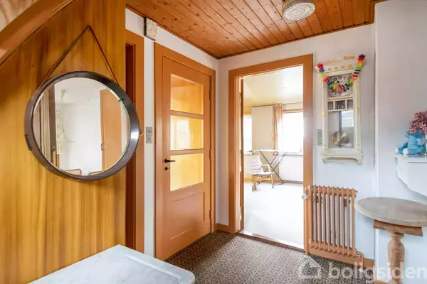 Et rundt spejl hænger på en trævæg i en gang. Dørene er orange, og en radiator er synlig. En åben dør leder ind til et rum med et strygebræt.