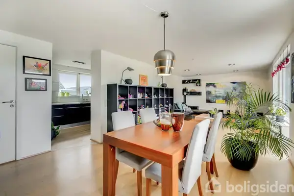 Et rektangulært træbord står i spisestuen med seks polstrede stole omkring. En moderne lampe hænger over bordet. Køkken ses til venstre, og stuen er i baggrunden.