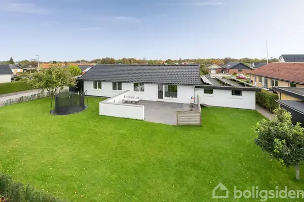 Huset ligger på en stor, frodig græsplæne med en terrasse foran. Ved siden af terrasseområdet er en trampolin. Omgivet af et kvarter med flere huse i baggrunden.