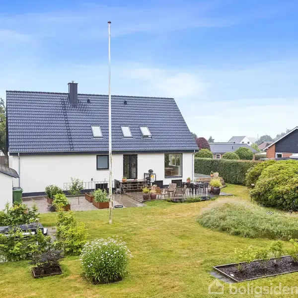 Hus med mørkt tag, stående i en velholdt baghave med græs, buske og terrasse. Omgivet af andre huse i et villakvarter under en blå himmel.