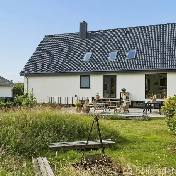 Hvidt hus med skråt tag, tre vinduer og terrassedør; i forhaven står et bord med stole. Omkring er en velplejet græsplæne og buske under en klar blå himmel.