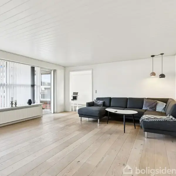 En mørkegrå hjørnesofa står i en lys stue med trægulve, ved siden af et rundt sofabord. Store vinduer fylder rummet med naturligt lys. Hylder og lamper pryder væggene.