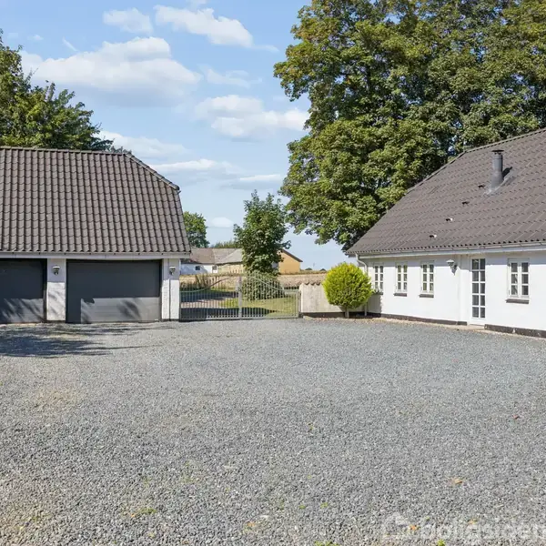 En hvid bygning med sort tag står til højre ved siden af en dobbelt garage. Grusbelagt gårdsplads omgivet af træer og en blå himmel.
