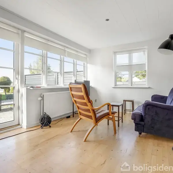 Stol og sofa står stille i en lys stue med store vinduer, der åbner ud mod en terrasse og have. Gulvlampen er slukket, og rummet er minimalistisk indrettet.