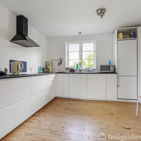 Et moderne køkken med hvide skabe, en indbygget ovn, og en emhætte. Et vindue kaster lys ind i rummet, hvor der også står en elkedel og en mikrobølgeovn. Lamineret trægulv.