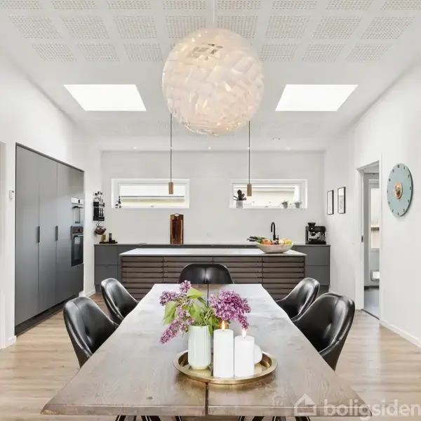 Spisebord med blomster og lys på en bakke; omgivet af sorte stole. Køkken med grå skabe i baggrunden, lys fra ovenlysvinduer. Moderne indretning med trægulv og stor loftslampe.