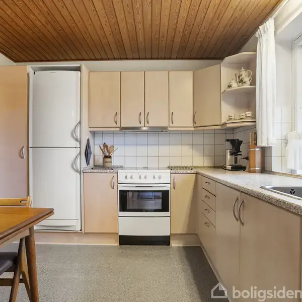 Køkkenet har en række beige skabe og skuffer, et hvidt køleskab og komfur. Der står køkkenredskaber og en kaffemaskine på bordet. Et vindue giver udsigt til haven.