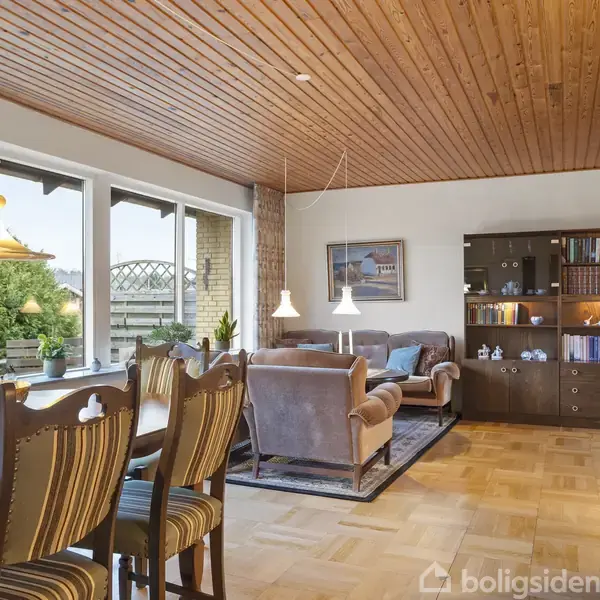 Stue med spisebord og stole. Sofaområde indeholder reol og lamper. Store vinduer giver udsigt til have. Træloftet og gulvet skaber en varm atmosfære.
