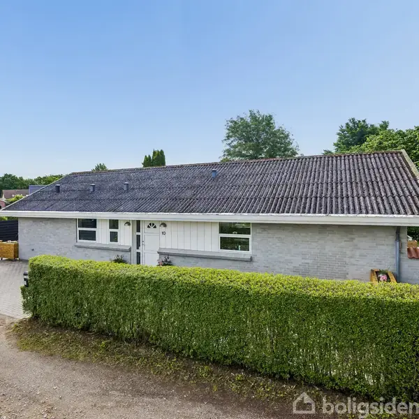 Hus står stille bag en tætrimmet hæk med grå murstensfacade og skråt, sort tag. En rød bil holder i indkørslen ved siden af huset i et grønt kvarter.