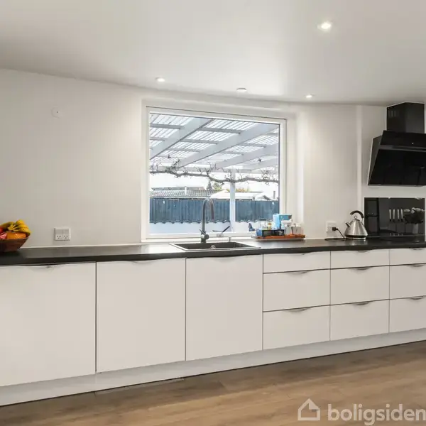 Moderne køkken med hvide skabe og sort bordplade. Køkkenet er rent, med en frugtskål og en vask med to armaturer. Store vinduer giver udsigt til en overdækket terrasse.