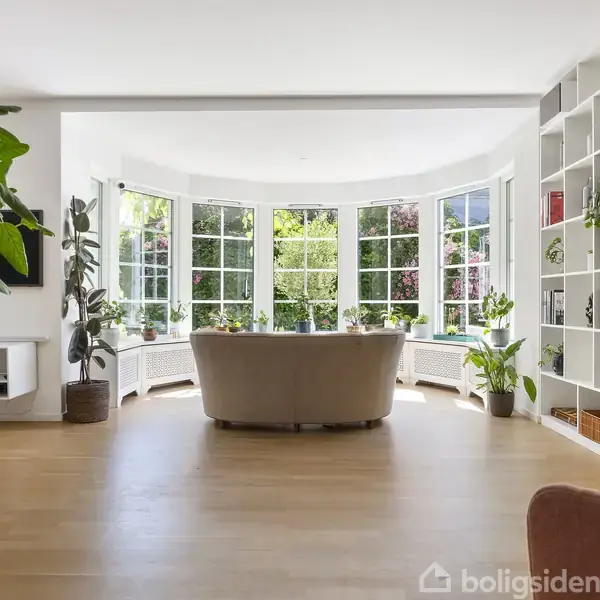 Stue med lys beige sofa foran store vinduer. Indbygget bogreol til højre, grønne planter spredt rundt i rummet. Lys træparketgulv, der skaber en lys og rummelig atmosfære.