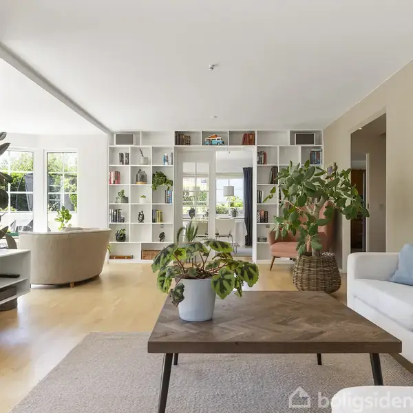 En moderne stue med et træbord i midten, omgivet af en hvid sofa og grønne planter. Store reoler fyldt med bøger danner en afslappet og lys atmosfære.