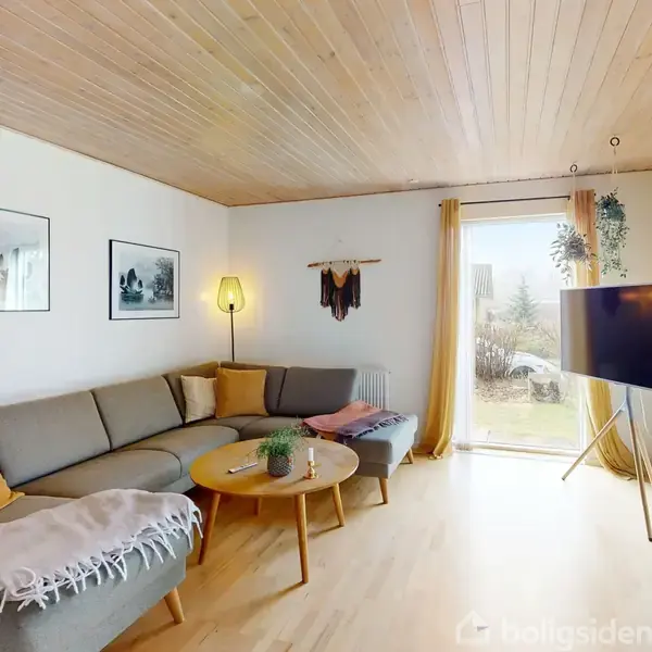 En beige hjørnesofa med puder står i et lyst rum med trægulv og træloft. En plante og et rundt sofabord med dekorationer er foran sofaen. Store vinduer med gardiner åbner til en have. En fladskærms-tv står på et stativ. To billedrammer hænger på væggen.