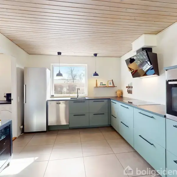 Moderne køkken med blå skabe, hvor en opvaskemaskine og ovn er integreret. Rummet er lyst med en central vask under et vindue, to hængende lamper og lyse fliser.