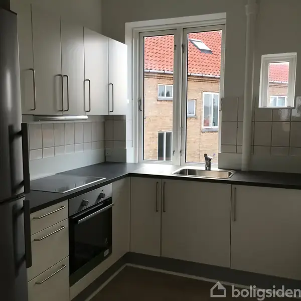 Et moderne køkken med hvide skabe, sorte bordplader og indbygget ovn. Et køleskab står til venstre, og store vinduer giver udsigt til murstensbygninger udenfor.