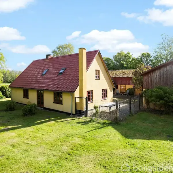 Et gult hus med rødt tag står på en grøn græsplæne. Omkring huset er træer og en mindre gårdbygning. Himmelen er klar med spredte skyer.