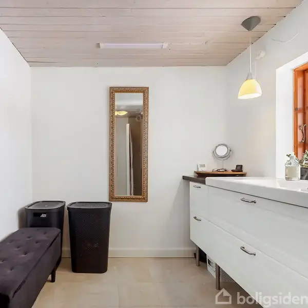 Badeværelse med hvidt skab og vask under vindue. Langt spejl hænger på væg, omgivet af to vasketøjskurve og en bænk. Loftlampe hænger over skabet.