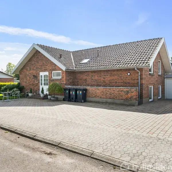 Rødt murstenshus med skråt tag står stille ved en flisebelagt indkørsel. To skraldespande og en garageport befinder sig på højre side. Roligt nabolag med grønne hække i baggrunden.