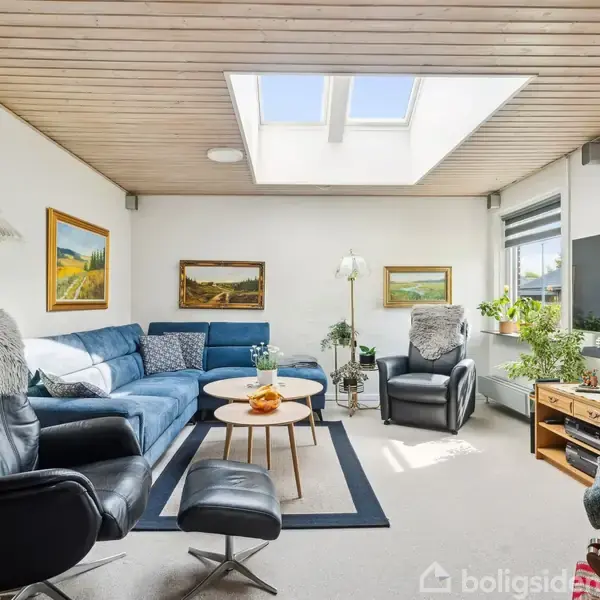 En stue med en blå hjørnesofa og sorte lænestole omkring et lavt, rundt sofabord. Ovenlys vindue giver naturligt lys, vægmalerier og en stor fladskærm på væggen.