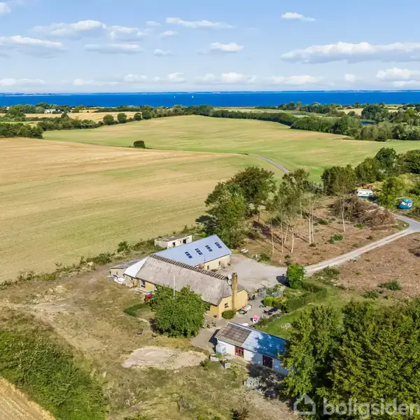 En gårdbygning står stille foran et stort åbent marklandskab med skov i horisonten og havet i det fjerne. Veje og træer omgiver ejendommen.