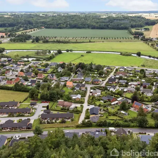 Byområde med huse spredt i et frodigt landskab; grønne marker og skov omkranser området. En vej snor sig gennem bebyggelsen, mens en flod flyder i baggrunden.