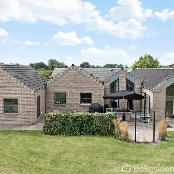 Moderne murstenshus står stille, omgivet af en velplejet græsplæne og få træer under en blå himmel med skyer. Huset har store vinduer og en terrasse med parasol.