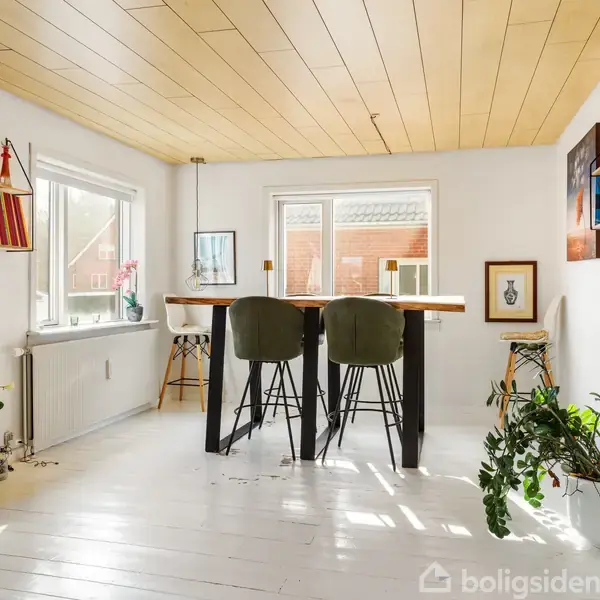 Et lyst rum med træbord og fire stole i midten. Store vinduer slipper sollys ind. Vægge har hylder med bøger og planter, og gulvet er hvidmalet.