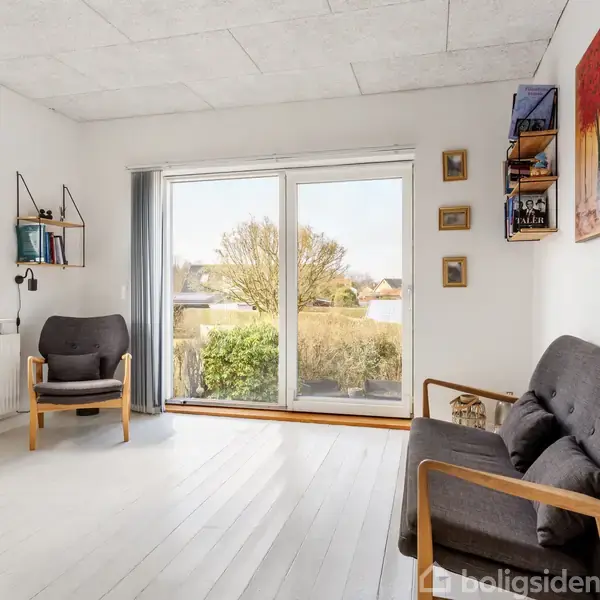 Stue med to grå lænestole, trægulv, store vinduer mod terrasse med udsigt til en have. Hylder med bøger og en maleri på væggen. Minimalistisk og lys indretning.