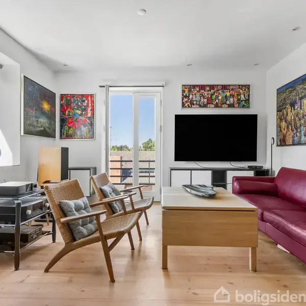 Sofaen er rød og står foran et stort TV. Stuen er lys med store malerier på væggene, træmøbler og et sofabord, omgivet af vinduer og en balkon.