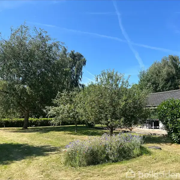 Træer står i en grøn have, mens blomsterbed blomstrer i forgrunden. En del af en terrasse med stole er synlig ved et mørkt tegltagshus. Klar himmel over.